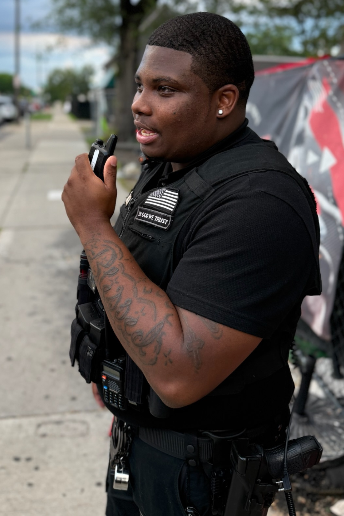 A police officer with tattoos on his arm, wearing a black uniform with patches and equipment, standing on a sidewalk and talking on a walkie-talkie.
