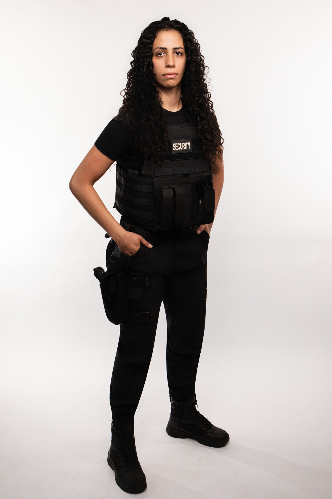 A woman in security uniform with a black vest labeled 'Security', black tactical pants, and black boots, standing with hands in pockets against a plain white background.