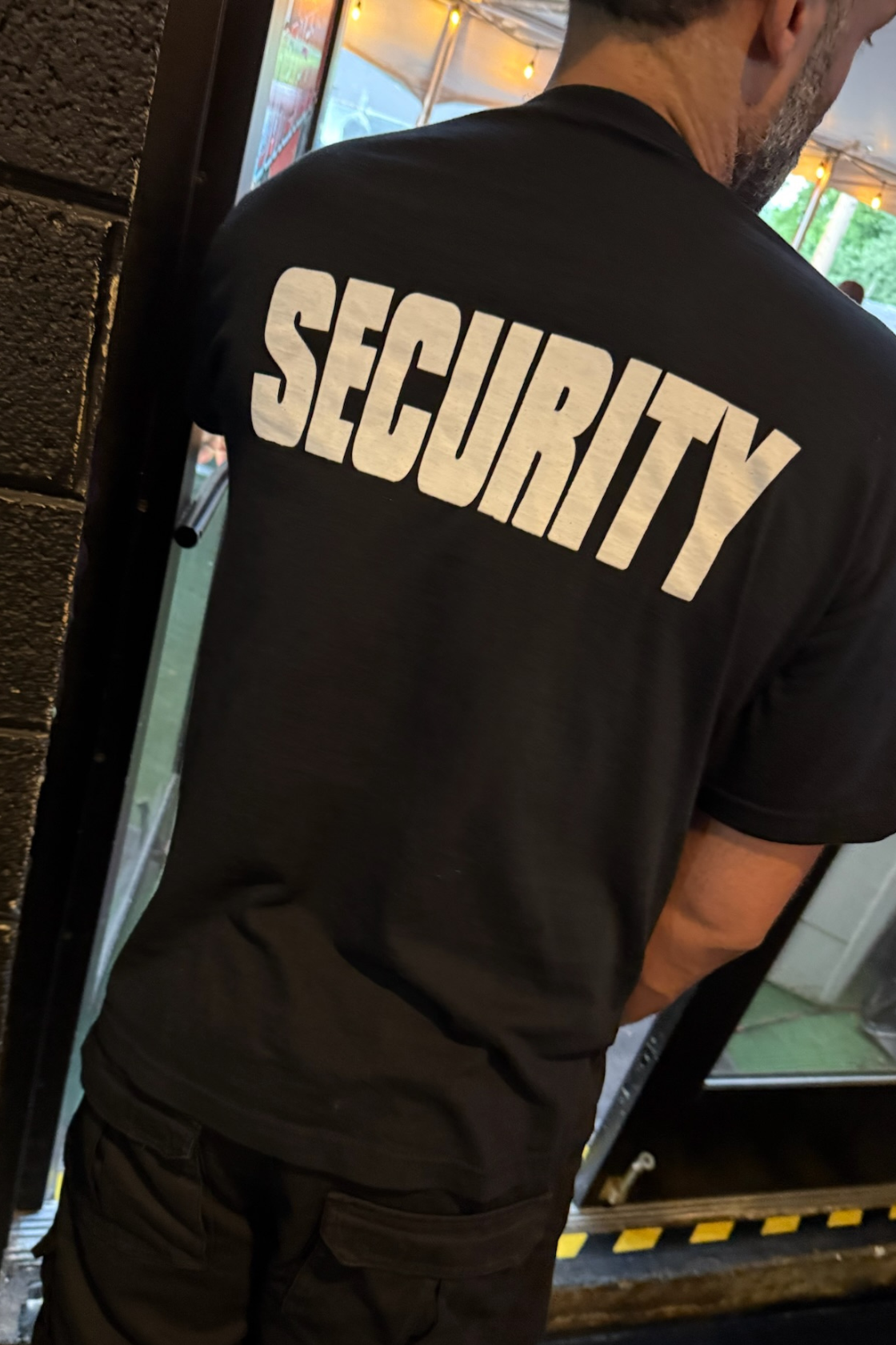A person wearing a black T-shirt with the word "SECURITY" printed on the back, standing near an entrance or exit door.