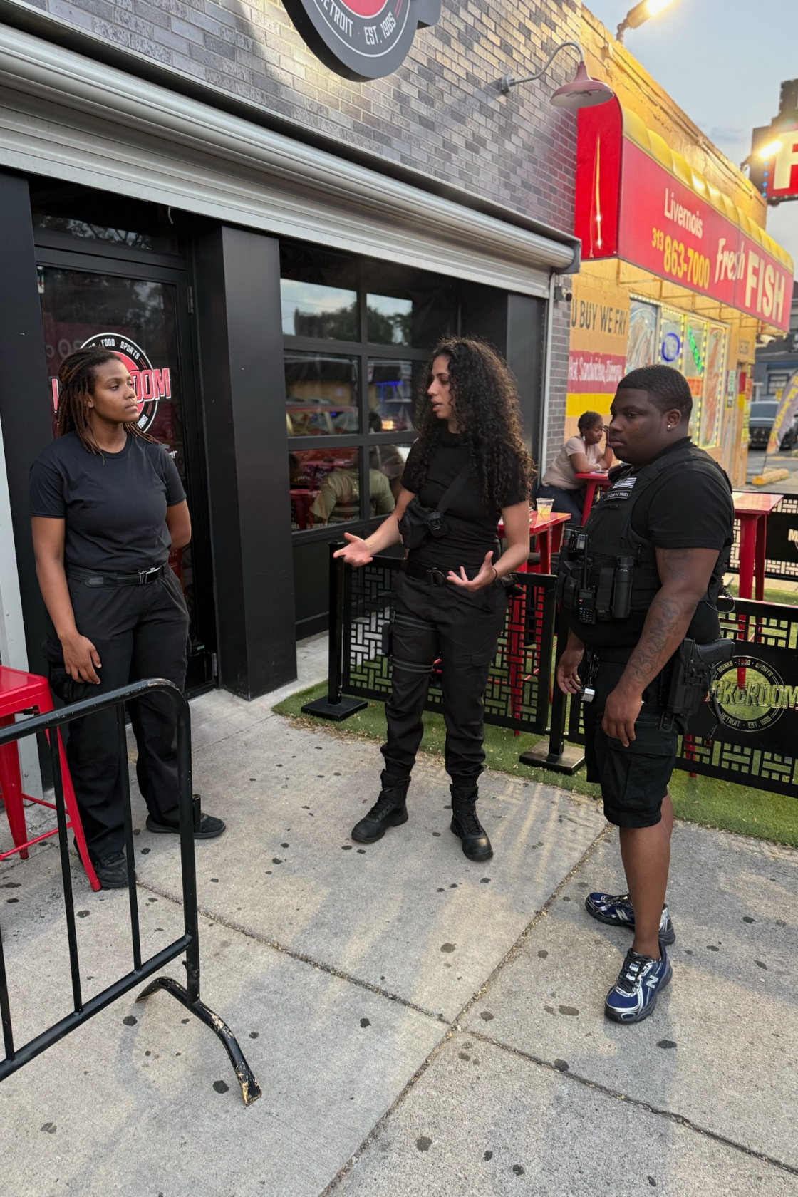Three people, two women and one man, stand outside a restaurant or food establishment on the sidewalk, engaged in a conversation, with a black security barrier nearby and a woman sitting at a red table in the background.
