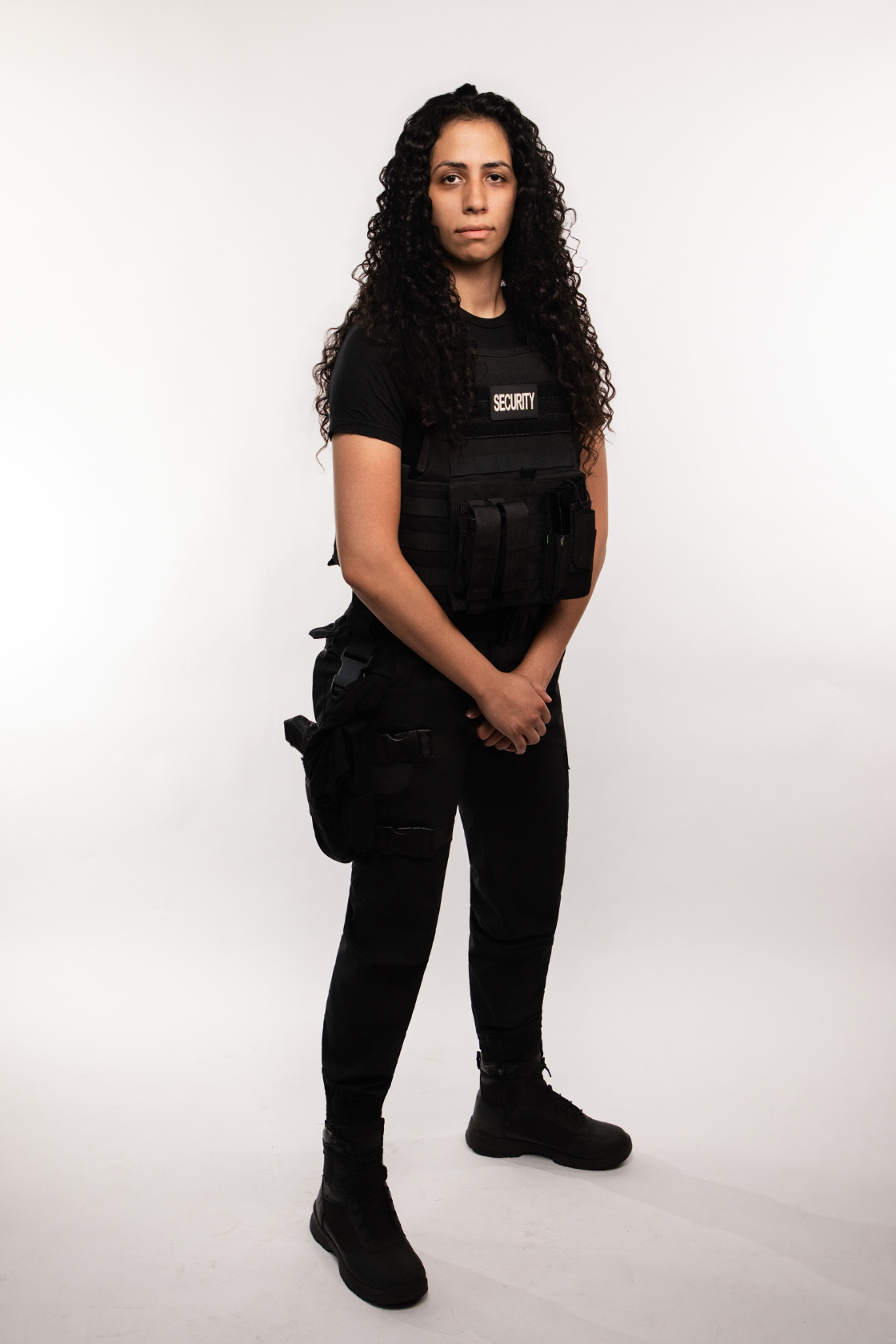A woman dressed in black tactical clothing with a security vest, standing confidently with hands clasped in front, against a plain white background.