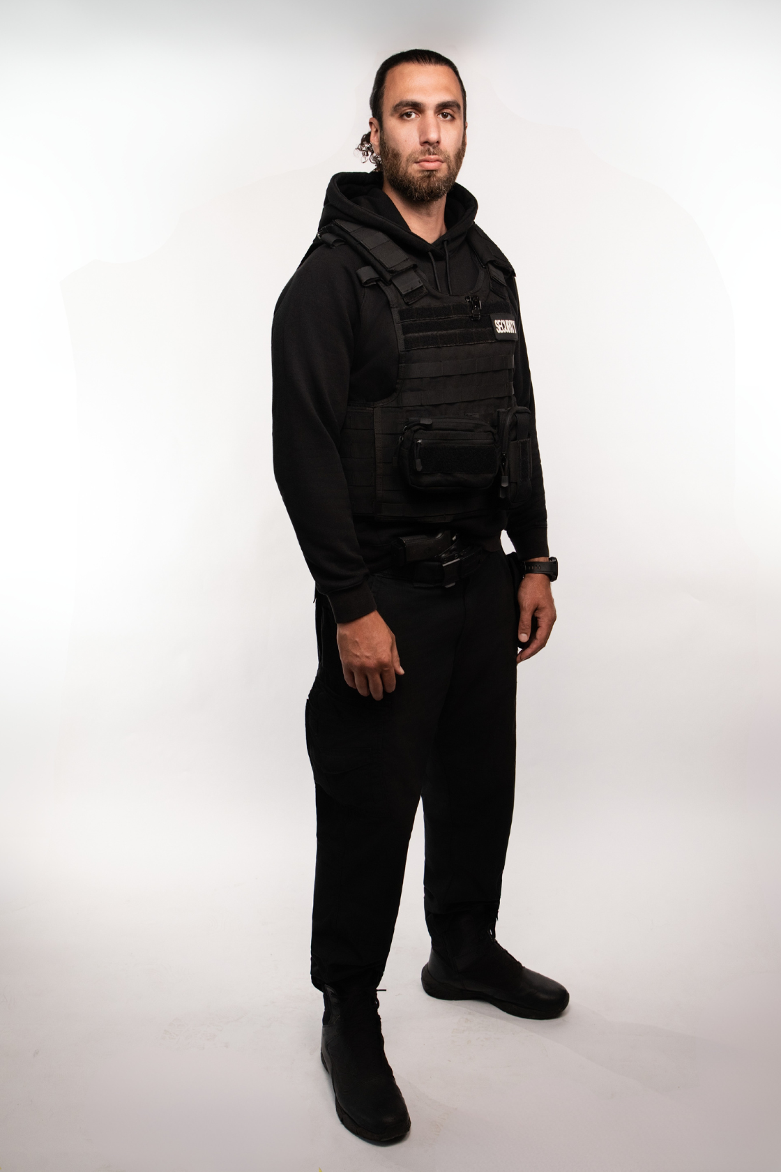 A male security guard standing against a plain white background, wearing a black uniform, tactical vest, and black boots.