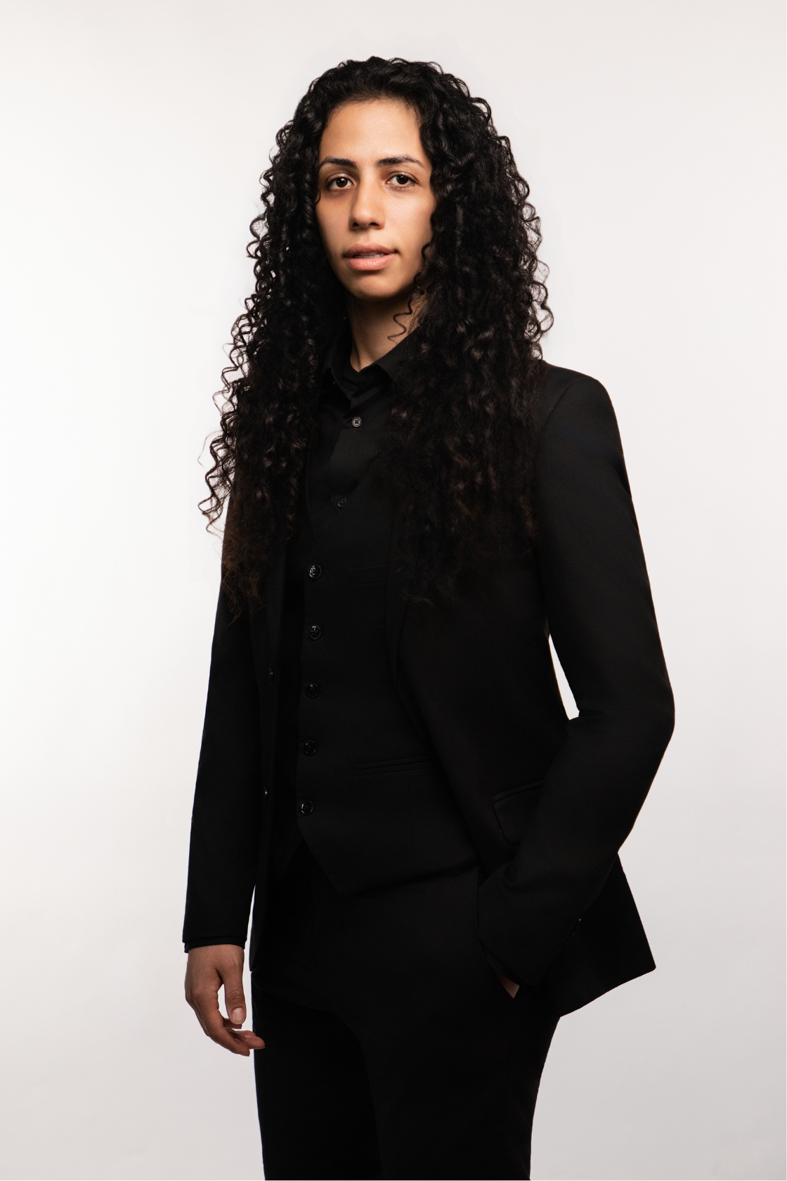A woman with curly dark hair wearing a black suit against a white background.
