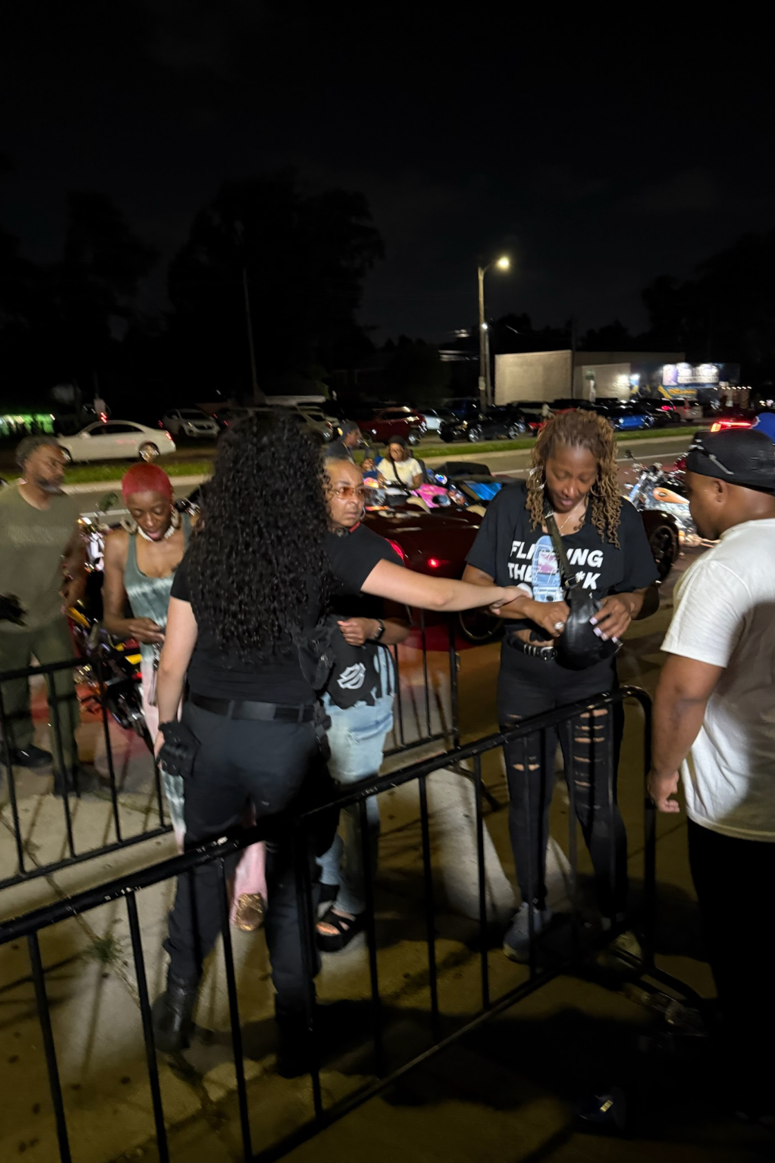 A group of people on a city street at night, with some standing near a black metal fence and others in the background, illuminated by streetlights and nearby cars. The scene appears casual, with some individuals exchanging items.