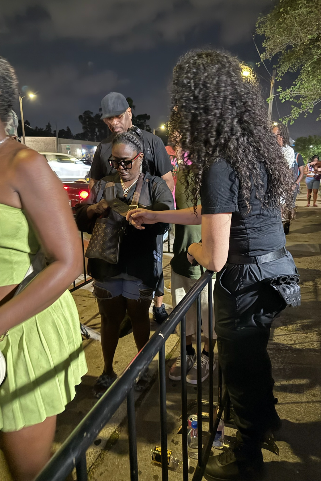 Group of people standing in line outdoors at night, some looking at their phones, with a black metal barrier in front of them, and cars and streetlights in the background.