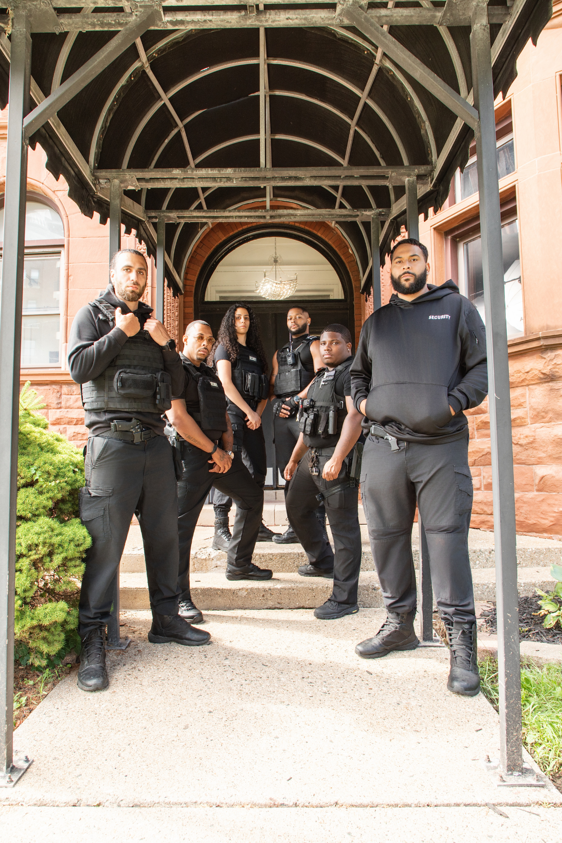 Group of six security officers standing on front steps of a building, wearing black tactical uniforms and gear.
