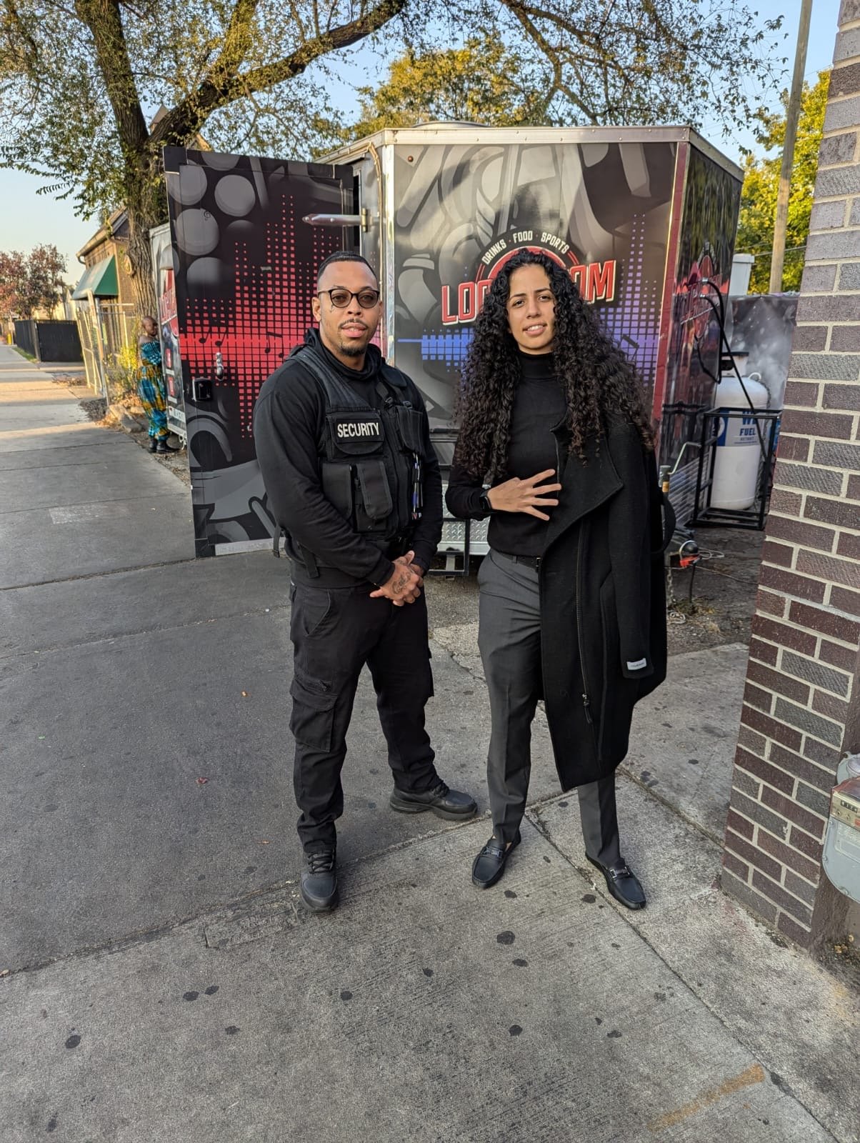 A security guard and a woman with long curly hair standing outdoors on a sidewalk in front of a food trailer with colorful graphics and the word 'LOOP' on it. There is a tree and a person in the background.