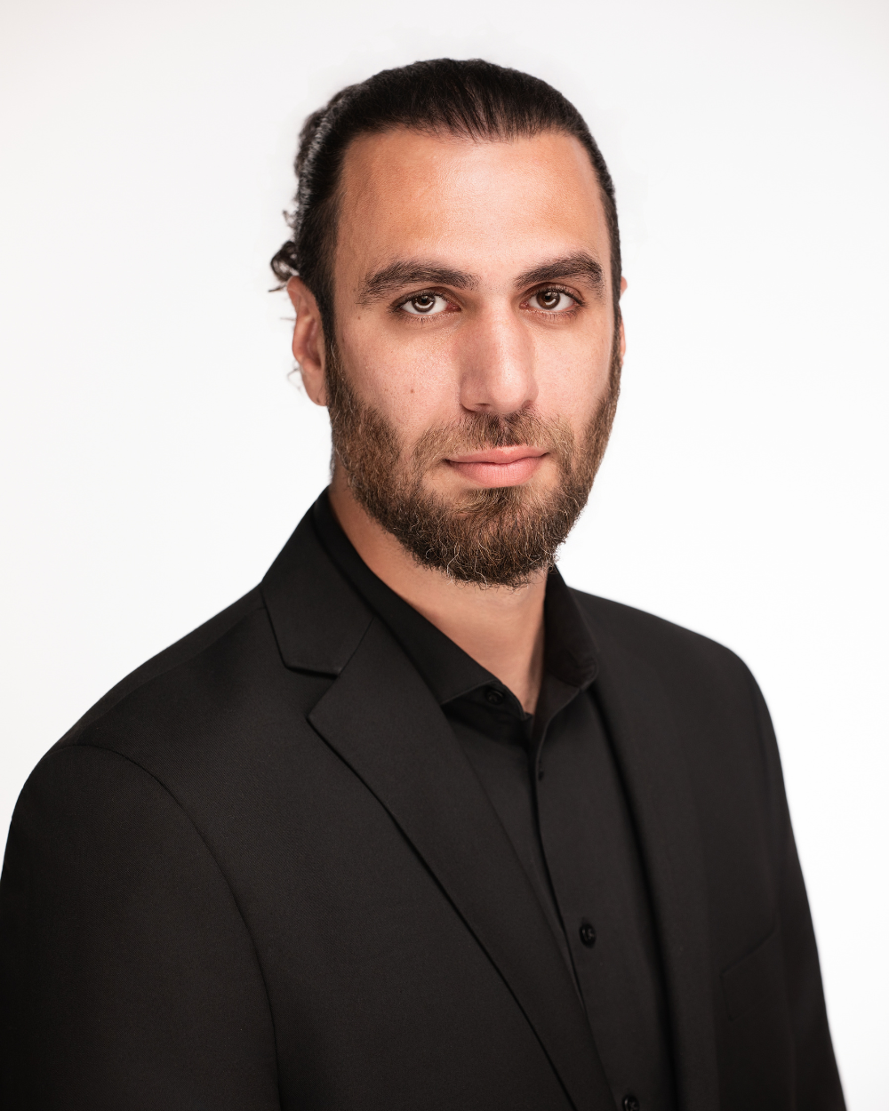 A man in a black suit standing indoors with wooden wall and plant in the background.
