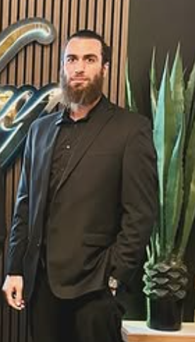 A man in a black suit standing indoors with wooden wall and plant in the background.