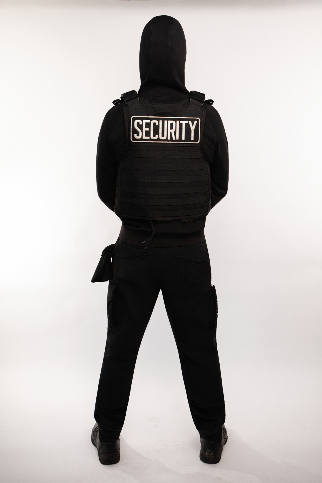 Person dressed in black security uniform with a vest labeled "SECURITY" on the back, facing away from the camera in front of a plain white background.