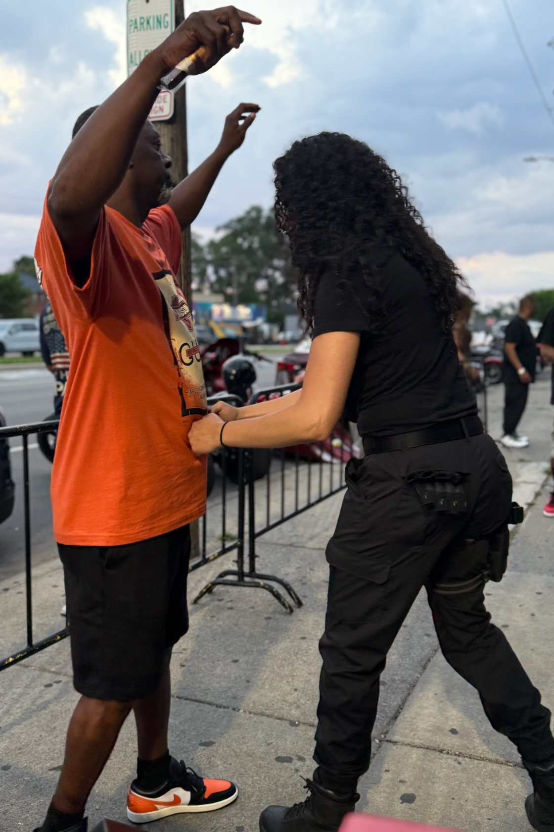 A woman in black pants and a woman in an orange shirt and black shorts are standing on a sidewalk, holding hands. The woman in black appears to be searching through her pocket or bag. There are other people and motorcycles in the background, with a c