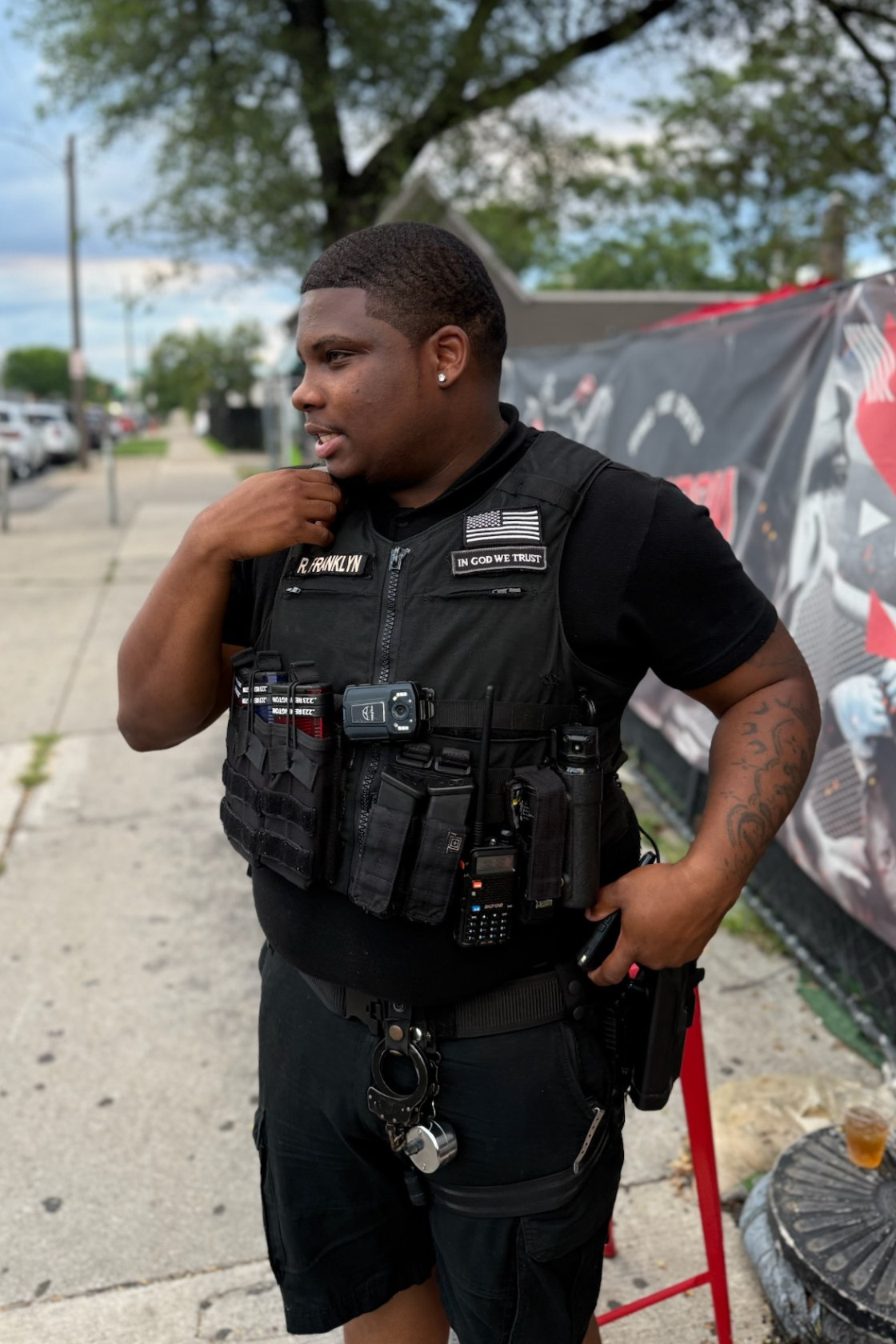 A young man wearing a black tactical vest with various communication and recording devices stands outdoors on a sidewalk, with trees and a street in the background.
