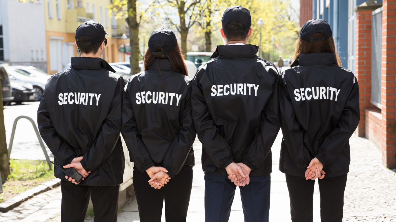 Four security guards in black jackets with 'SECURITY' written on the back, standing with their backs to the camera on a city sidewalk with trees and buildings in the background.