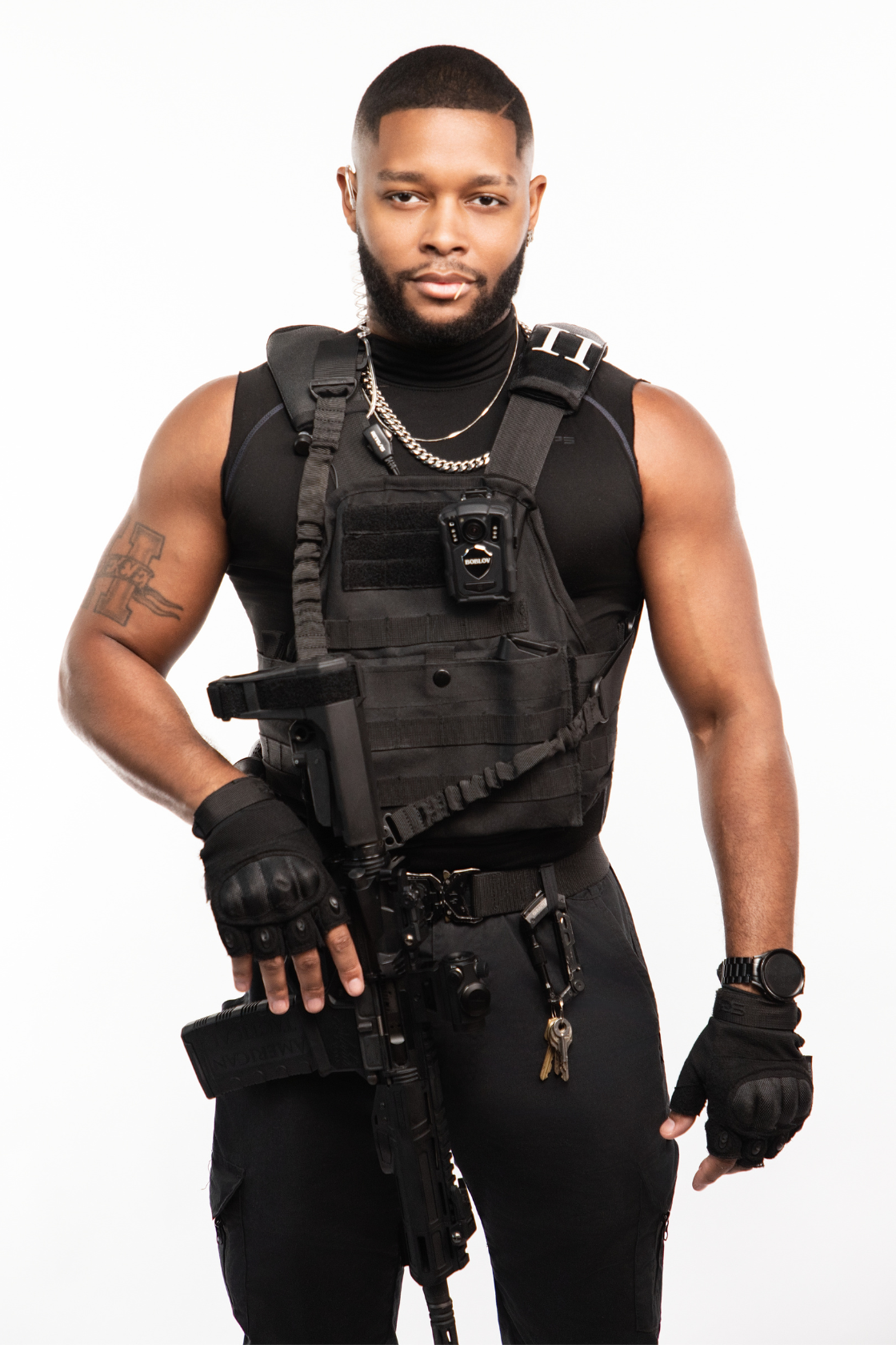 A man with a beard and short hair wearing tactical gear, including a vest with a radio and a gun, gloves, and a watch, standing against a white background.