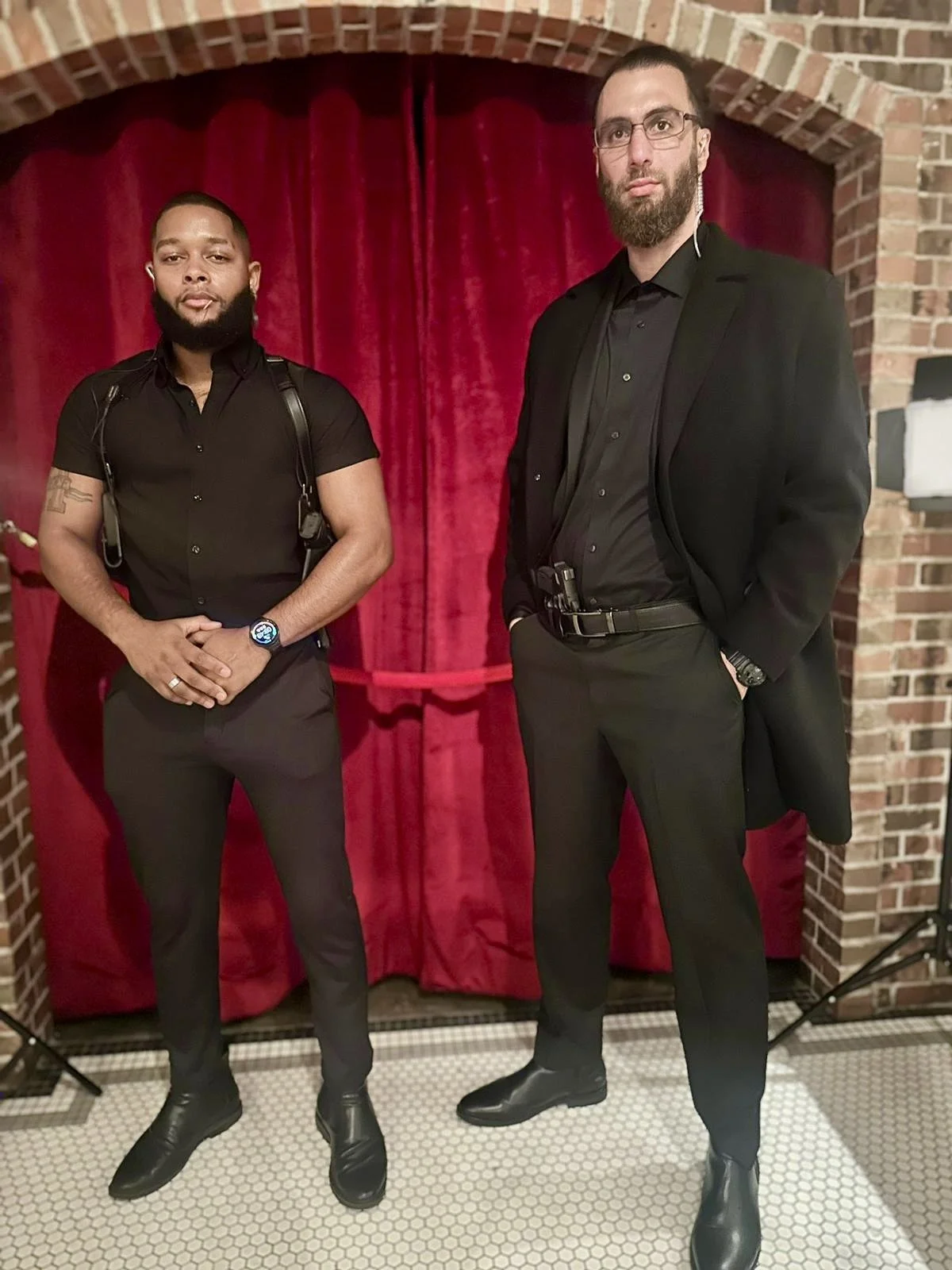 Two men dressed in black suits standing in front of a red curtain with brick walls on either side.