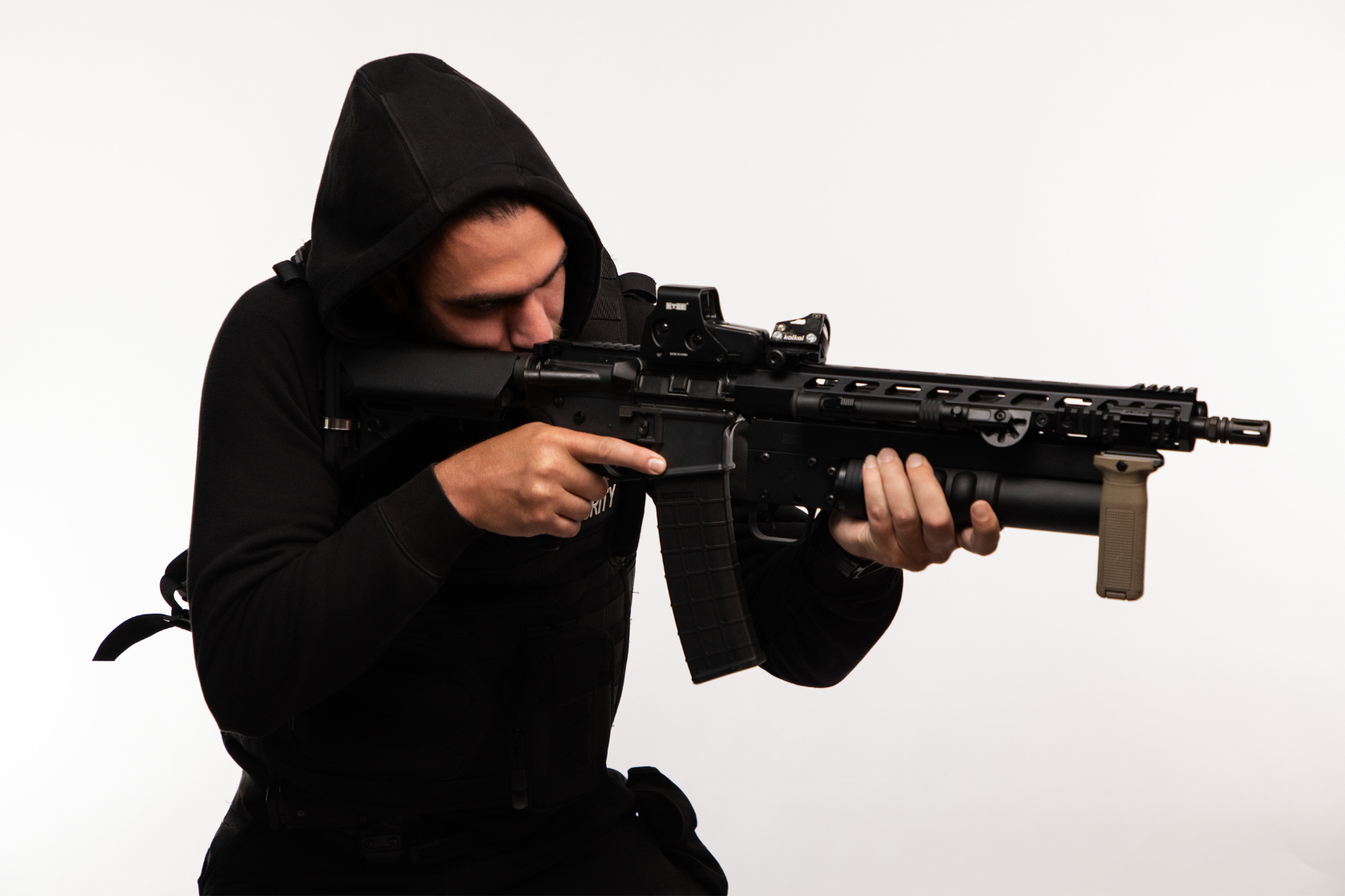 A person wearing a black hoodie and tactical gear aiming a black rifle with a scope in front of a plain white background.