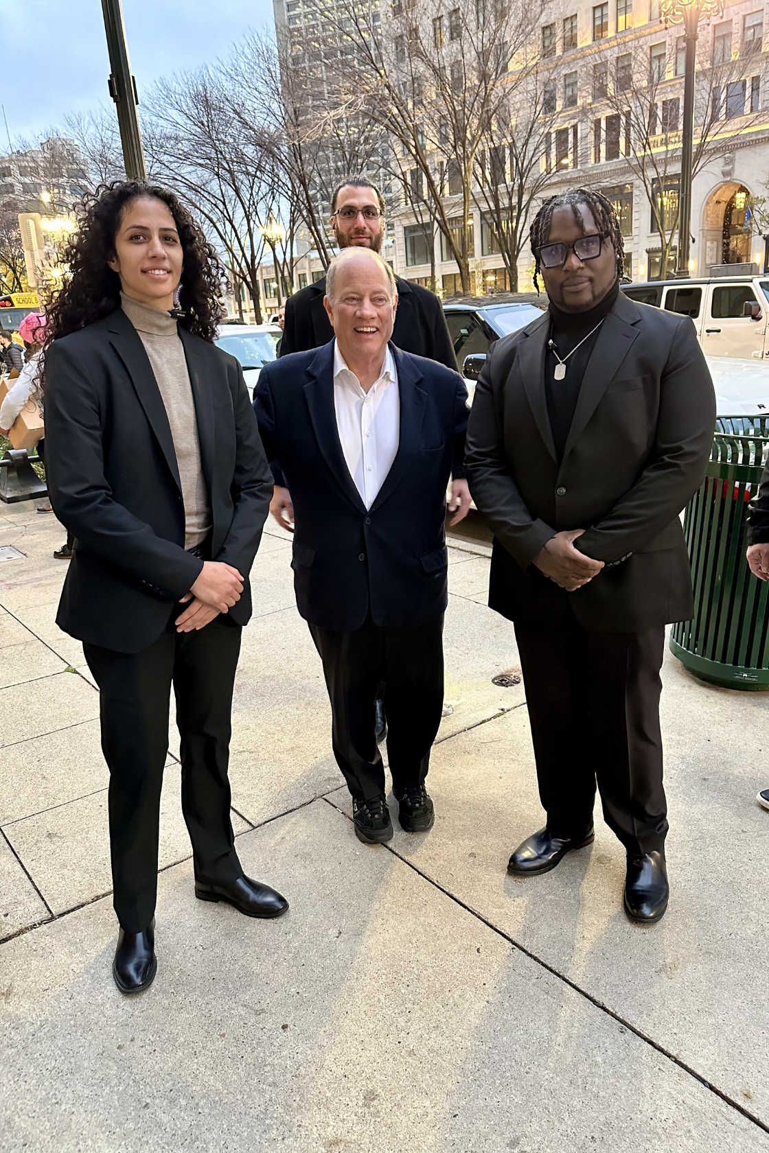 Four individuals in formal attire standing on a city sidewalk in front of leafless trees and tall buildings during sunset.
