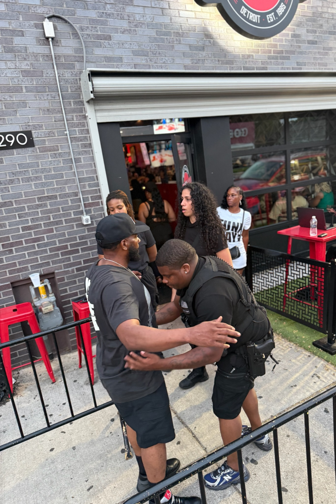 Two men, one in a black t-shirt and the other in a black security uniform, are interacting outside a building with black brick walls. Several women are standing nearby, and there are red chairs, a water bottle, and a black and red fence.