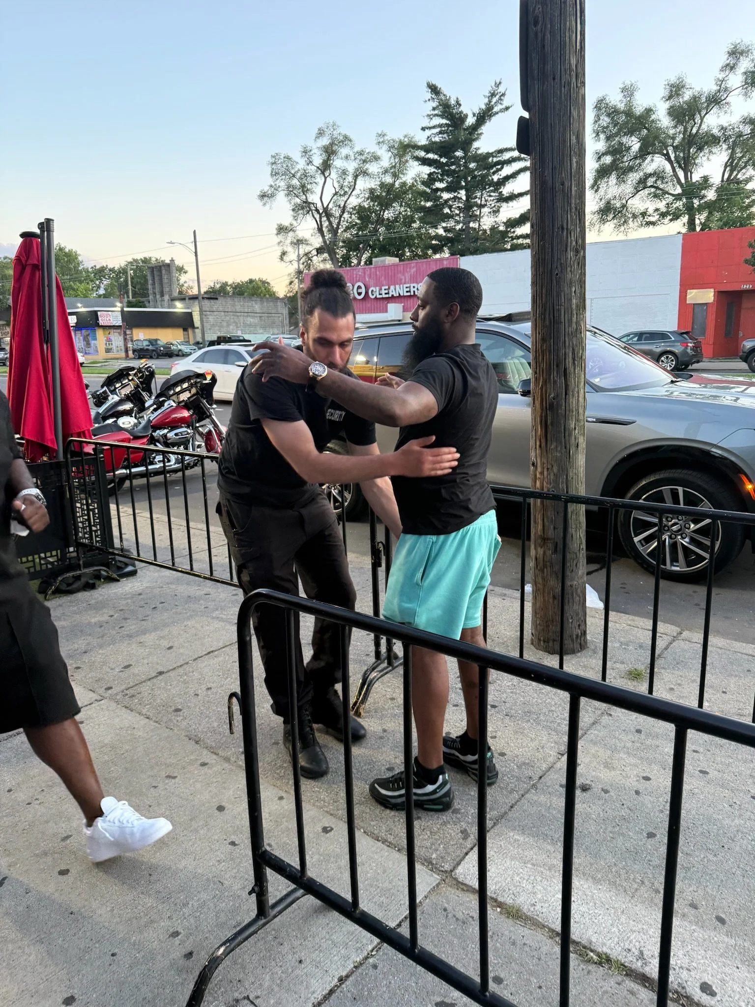 Two men are standing on a sidewalk near parked cars and motorcycles, engaged in conversation. One has long hair tied up, wearing a black shirt, and the other has short hair, a beard, and is wearing a black shirt and turquoise shorts. There is a red u
