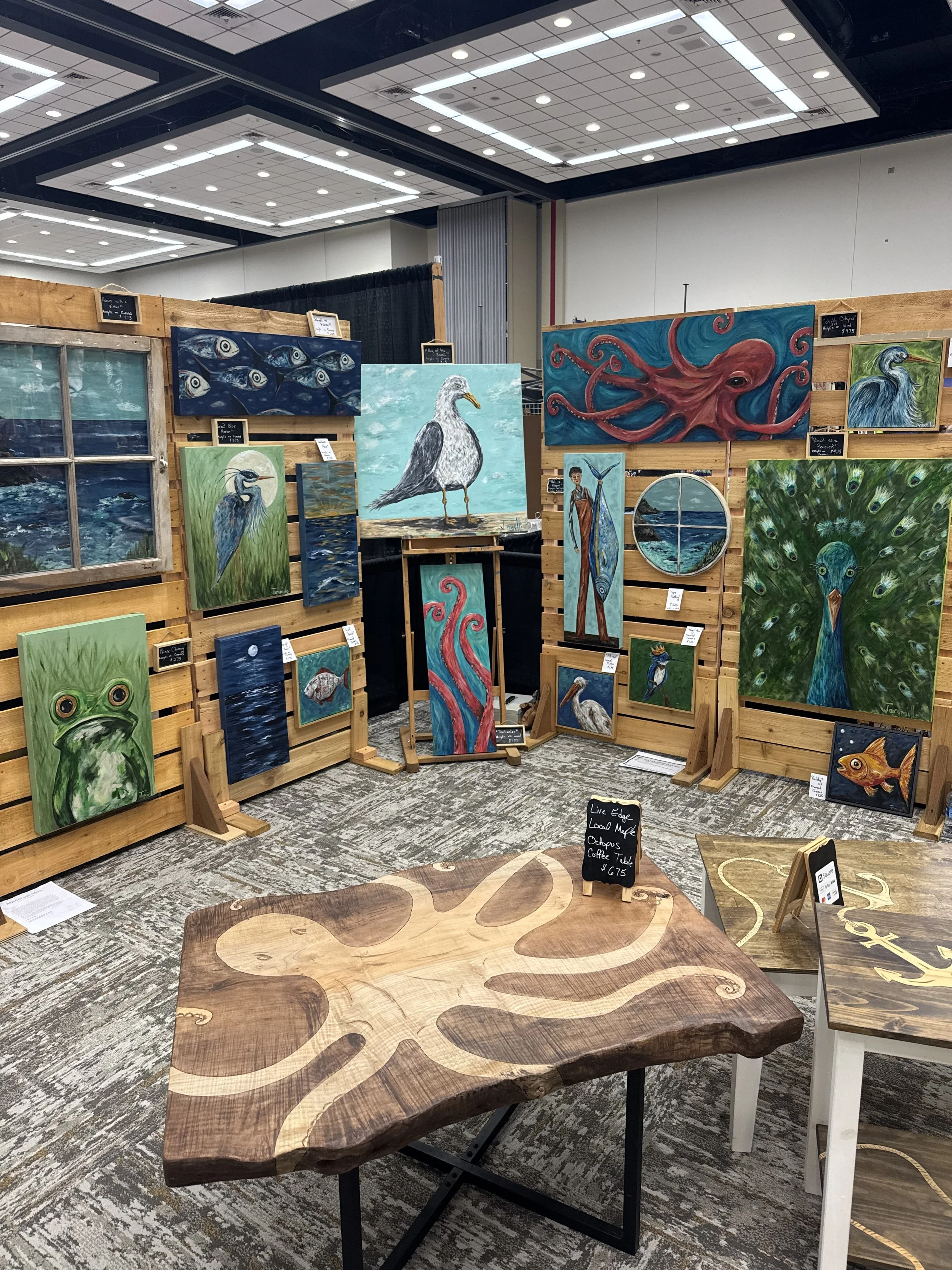 Wooden tables with natural, live edge finish are on display at an art fair, with paintings of animals and nature scenes hanging behind them.