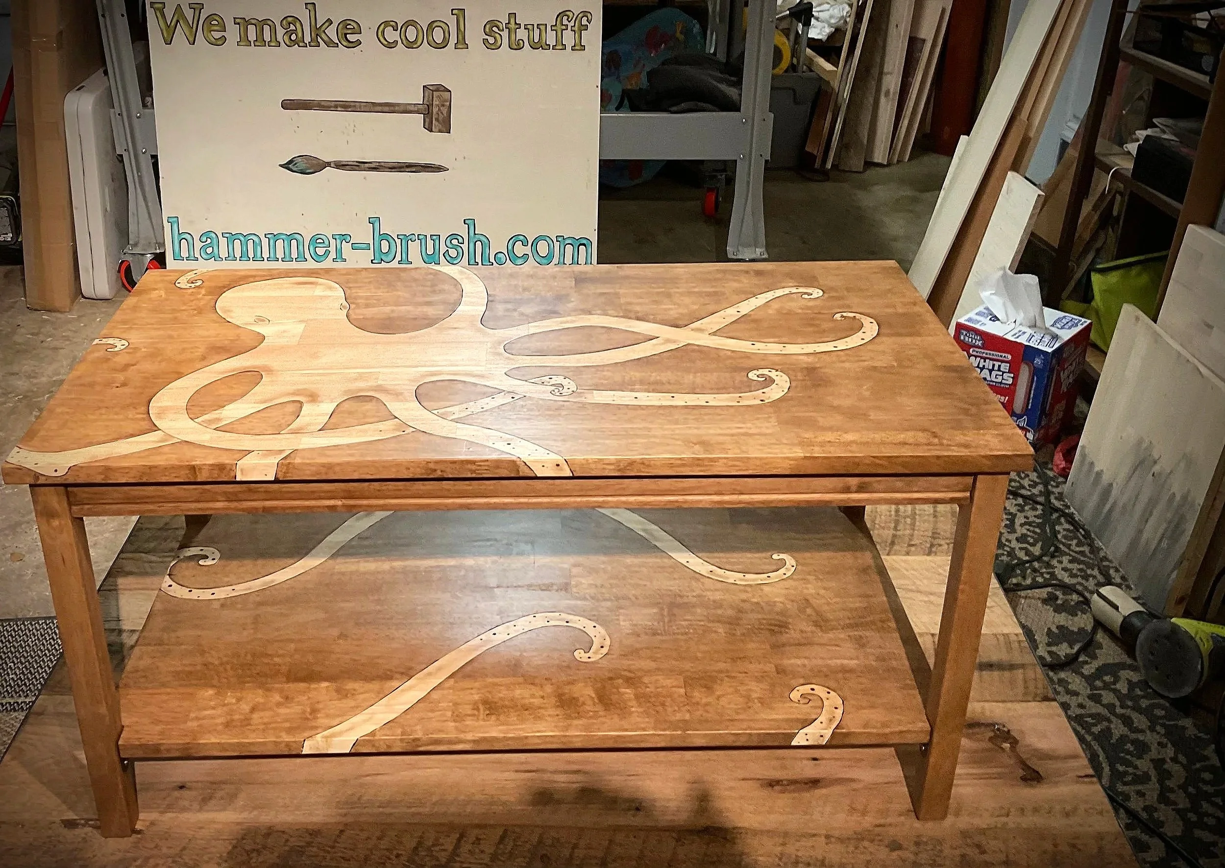 Wooden table with a carved octopus design on the surface, in a workshop setting. A sign in the background reads "We make cool stuff" and displays a hammer and chisel, with a website address hammer-brush.com.