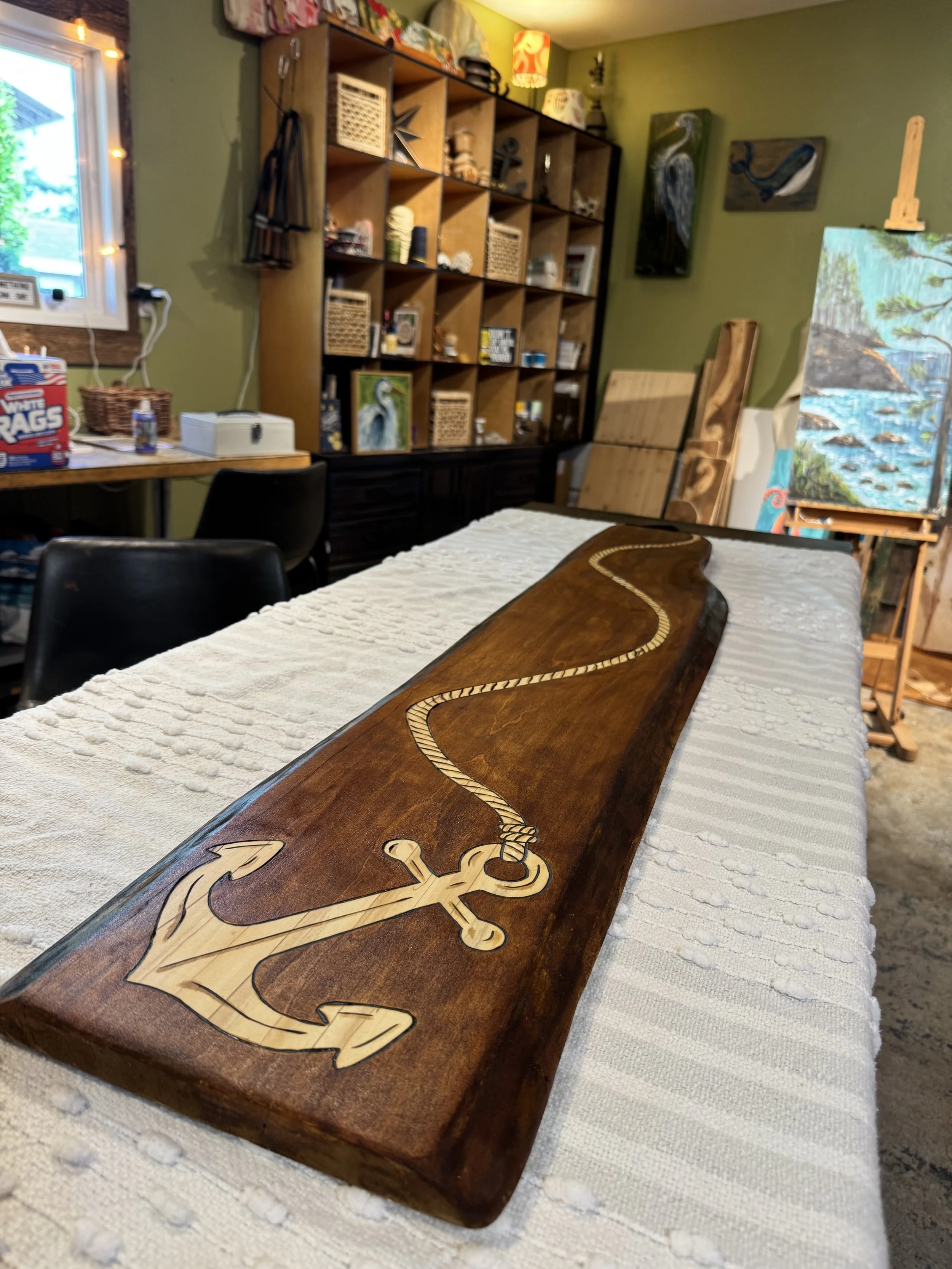 A wooden anchor-shaped decorative piece with a rope knot design, placed on a white tablecloth. The background shows a room with green walls, a black shelving unit filled with various items, and canvas paintings of herons and seagulls.