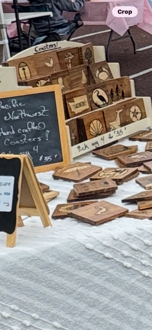 Handmade wooden coasters with nature and animal designs displayed on a table at a market stall. A chalkboard sign indicates prices, and there is a small wooden rack holding some of the coasters. Pink tablecloths and chairs are visible in the background.