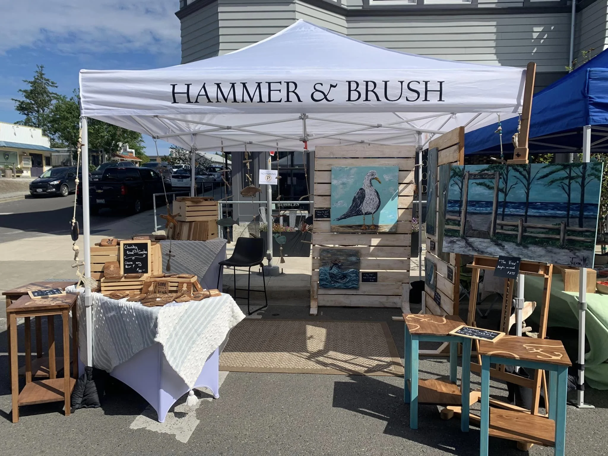 Outdoor art market booth with paintings of birds and trees, small wooden tables displaying jewelry or small art pieces, under a white tent with sign 'Hammer & Brush'
