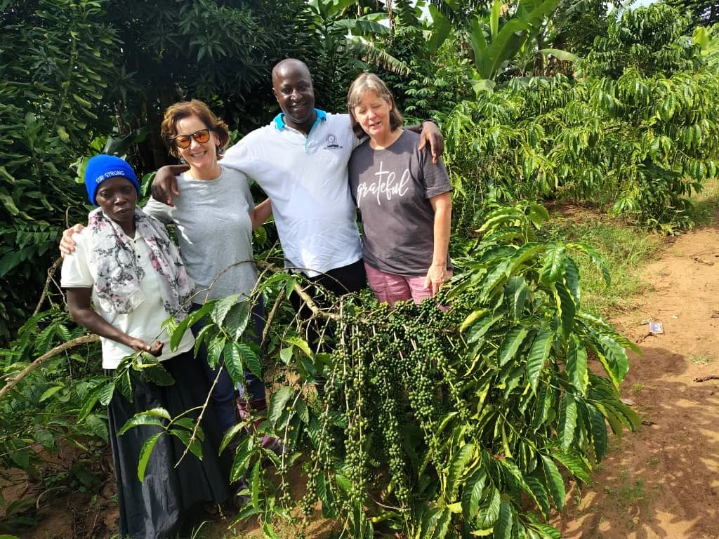 Four people standing together outdoors, smiling, with coffee plants in a lush green plantation.