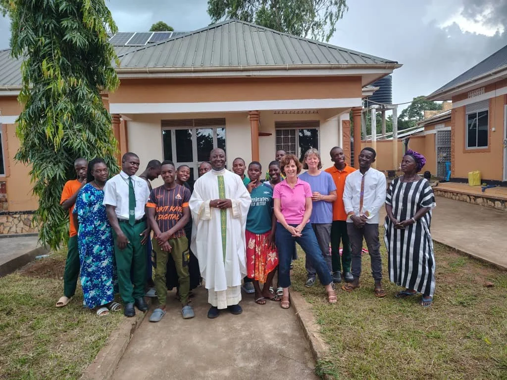 Group of people standing outside a house, including men and women, some in traditional and casual clothing, with a priest or leader in white robes in the center, and a tree on the left.
