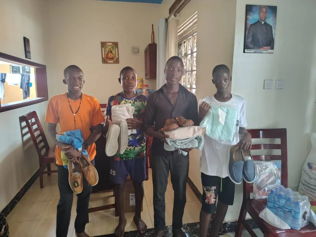 Four boys standing indoors, holding shoes and folded clothes, with water bottles and supplies on a chair to the side.