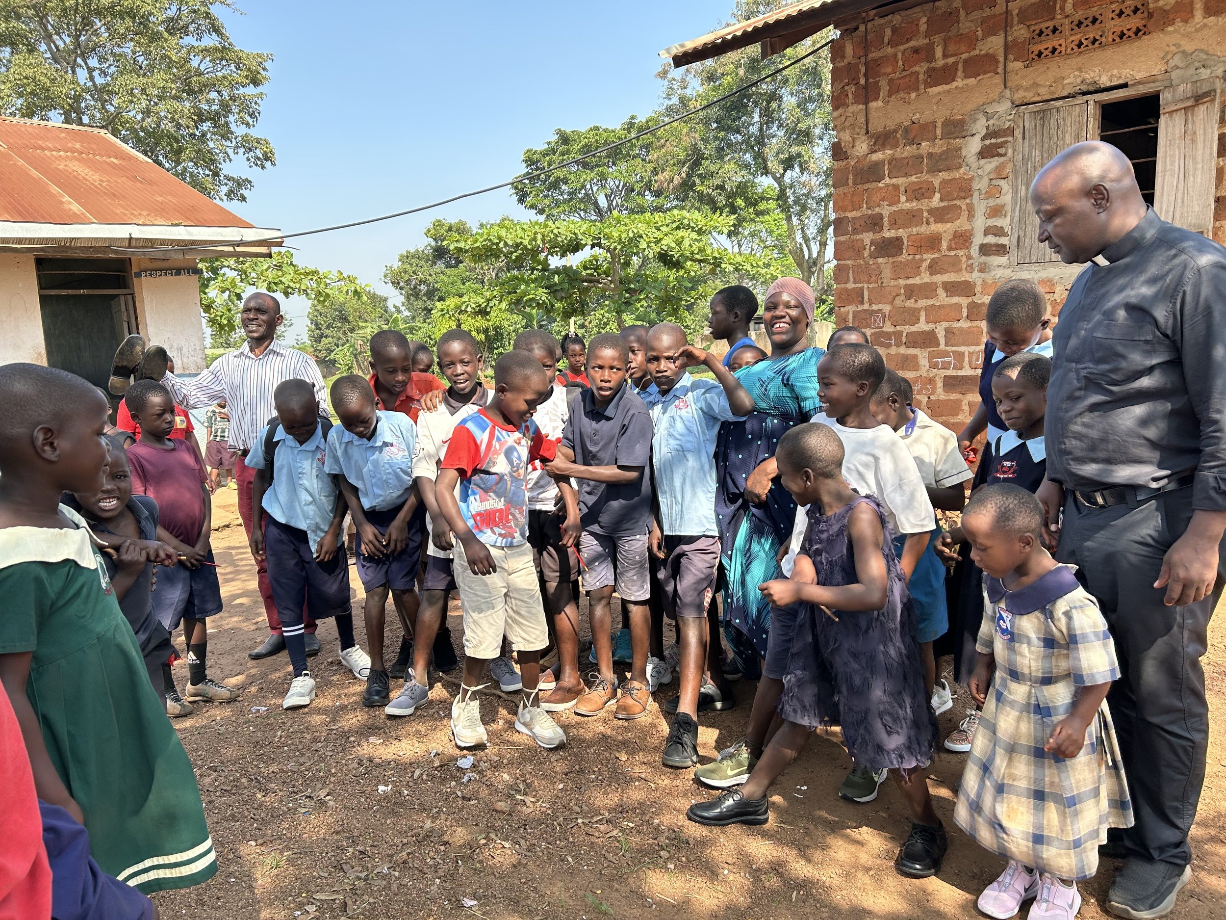 A group of children and a man, possibly a teacher or community leader, gathered outdoors in a small village setting with brick and plaster houses and lush green trees, engaging in a communal activity or discussion.