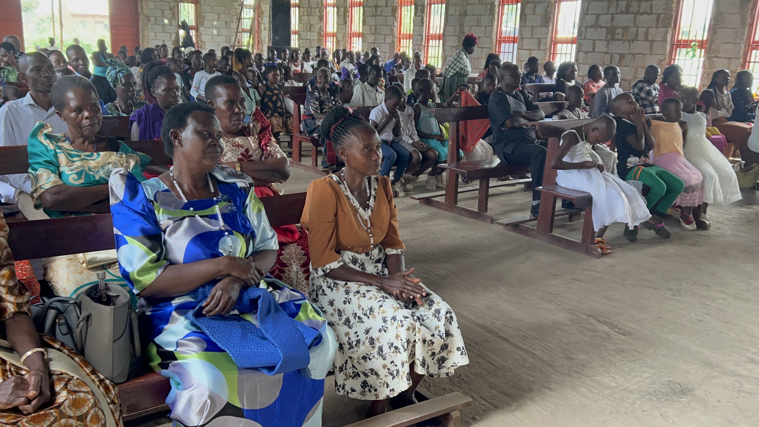A large group of people, including women and children, sitting on benches inside a partially constructed building with brick walls and large windows, attending a community gathering or event.