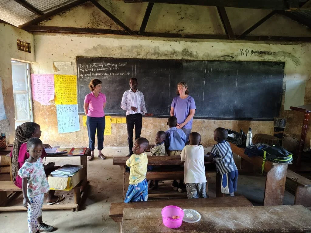 A classroom with children and three teachers or adults. Children are seated at wooden desks and standing around, while the adults stand at the blackboard, which has some writing. The classroom has a corrugated metal roof, worn walls, and colorful charts on the walls.