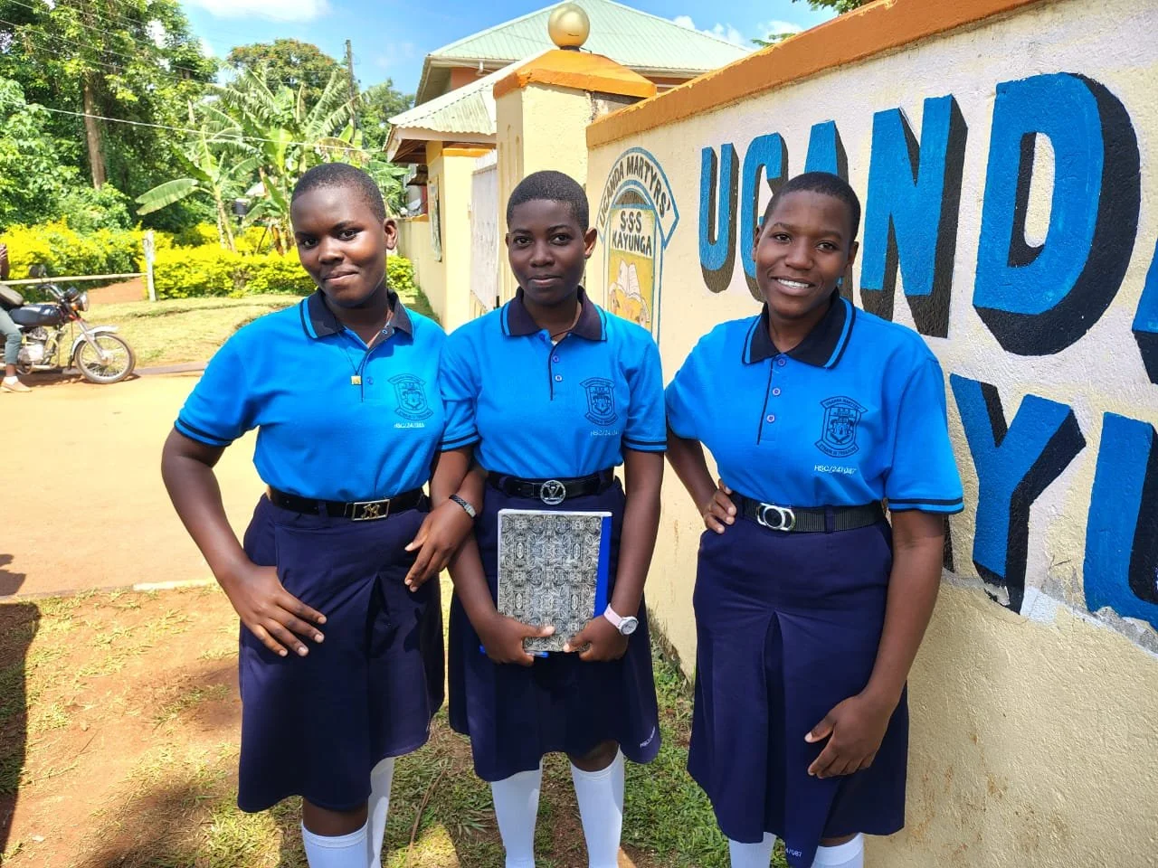 Three schoolgirls in blue uniforms standing outside near a yellow wall with blue text, smiling at the camera, one holding a notebook, in a green outdoor area.
