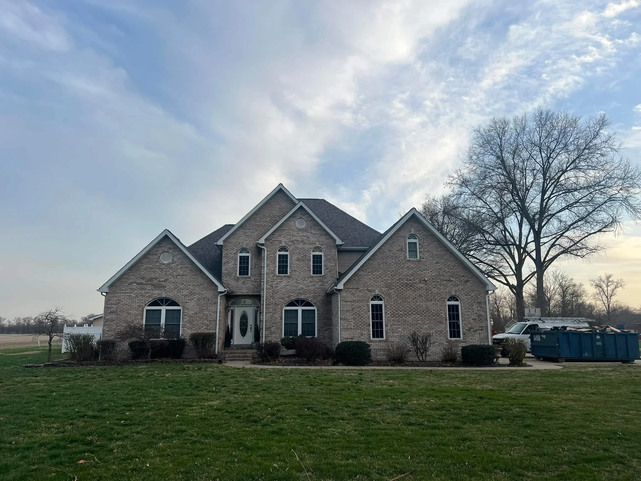 A two-story brick house with arched windows and a peaked roof is situated on a lawn with bushes and trees. A large, leafless tree stands to the right of the house. The sky above is partly cloudy with patches of blue.