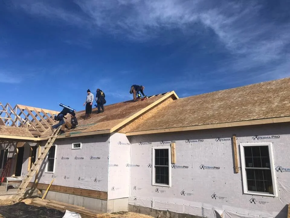 Construction workers installing a new roof on a house during daylight, with some workers on the roof and others on scaffolding, under a partly cloudy sky.