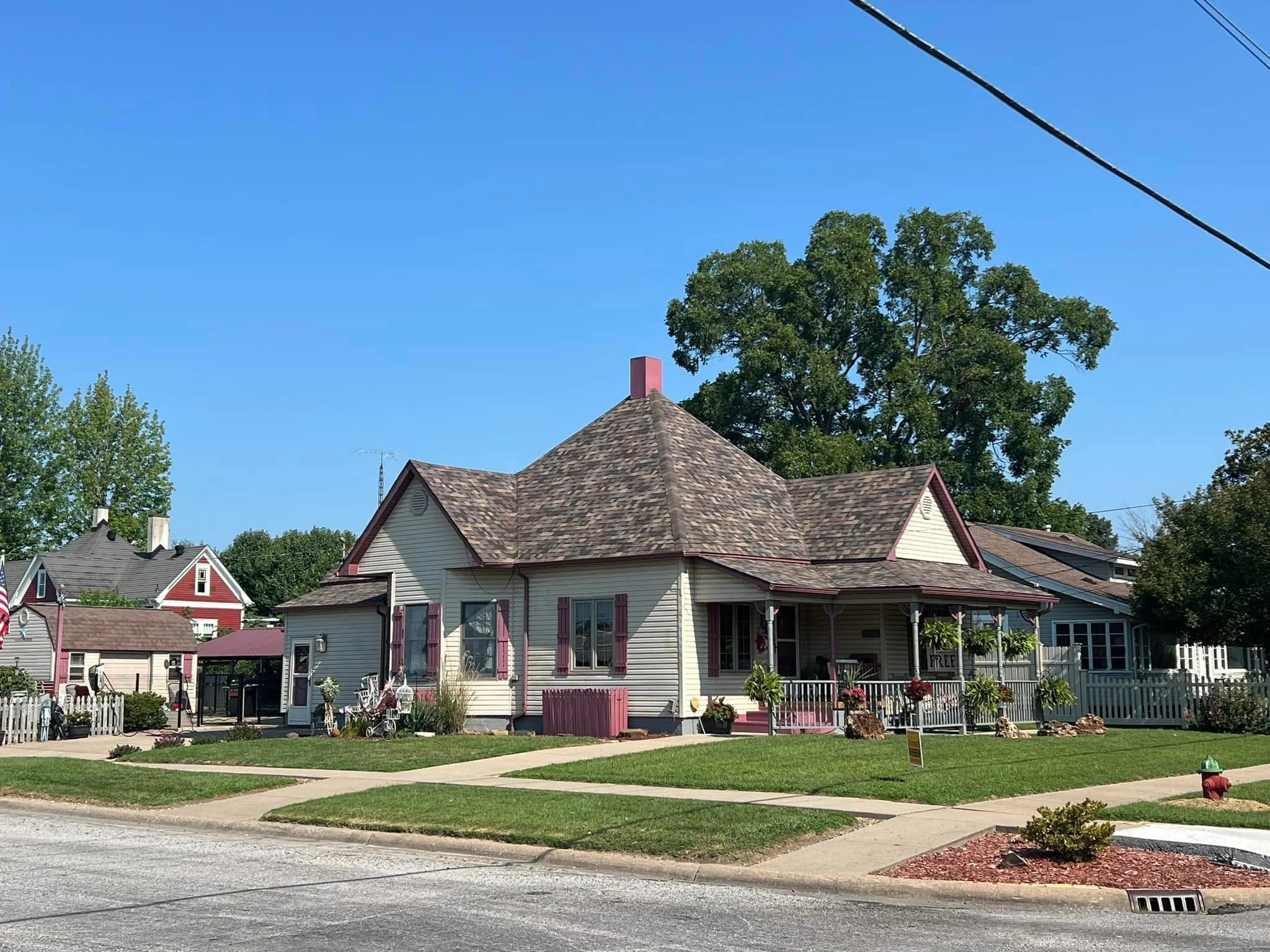 A quaint house with a steep gable roof, painted cream with red trim, and surrounded by a white picket fence. The front yard has a neatly maintained lawn, flower beds, and garden decorations. There are large trees and neighboring houses in the background under a clear blue sky.