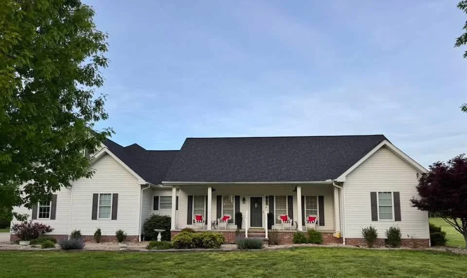 Single-story white house with a black roof, front porch with four columns, and holiday decorations such as red bows and small trees in front, surrounded by a green lawn and trees.