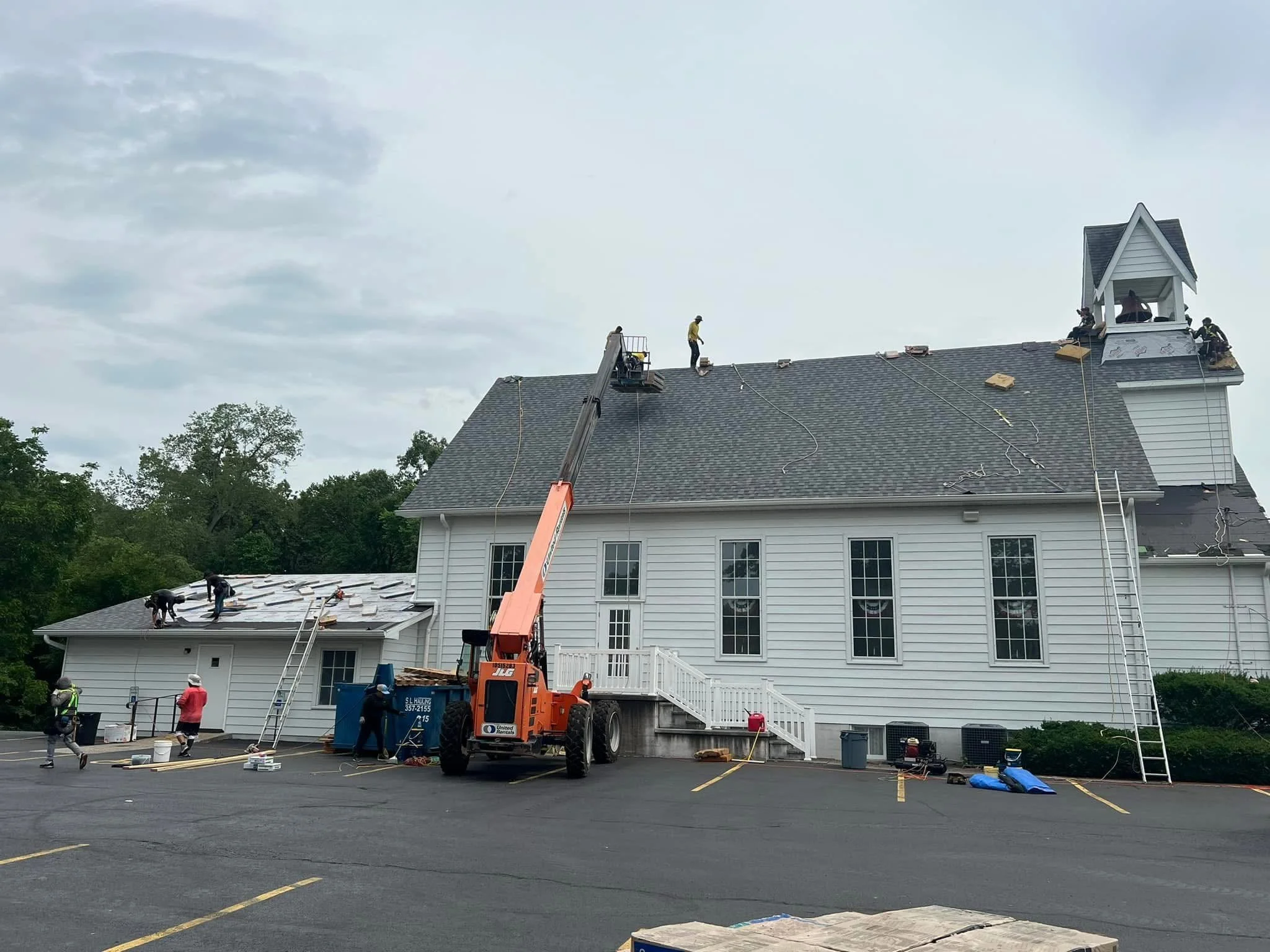 Workers installing new shingles on the rooftop of a white two-story building with a small tower. Construction equipment and tools are visible, and workers are on the roof and ground.