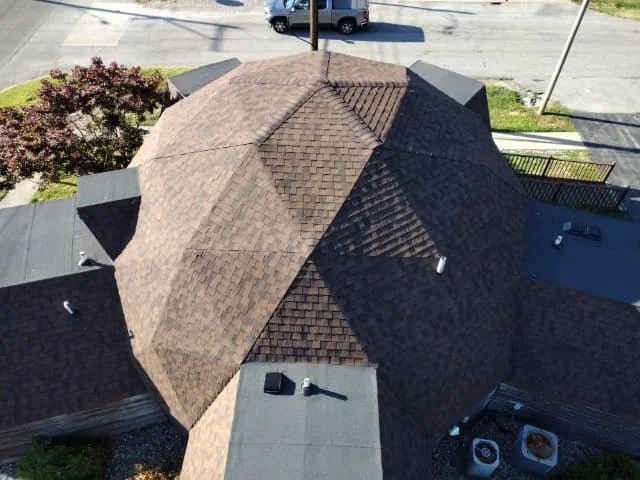 Aerial view of a house with a large, complex brown shingle roof, surrounded by trees, a driveway, and neighboring houses.