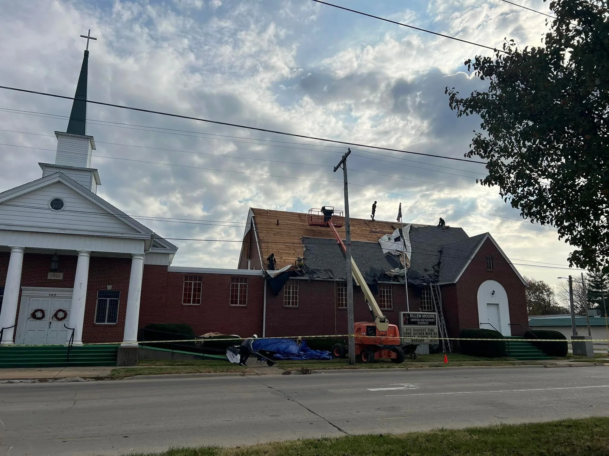 A church with a tall steeple and a cross on top, under a partly cloudy sky. Construction workers are working on the roof of a nearby building, with construction equipment and barriers visible in the foreground.