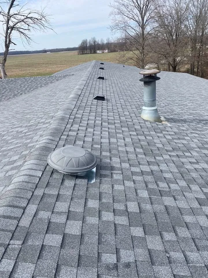View of a gray asphalt shingle roof with vents and a chimney pipe, overlooking a rural landscape with fields and bare trees.
