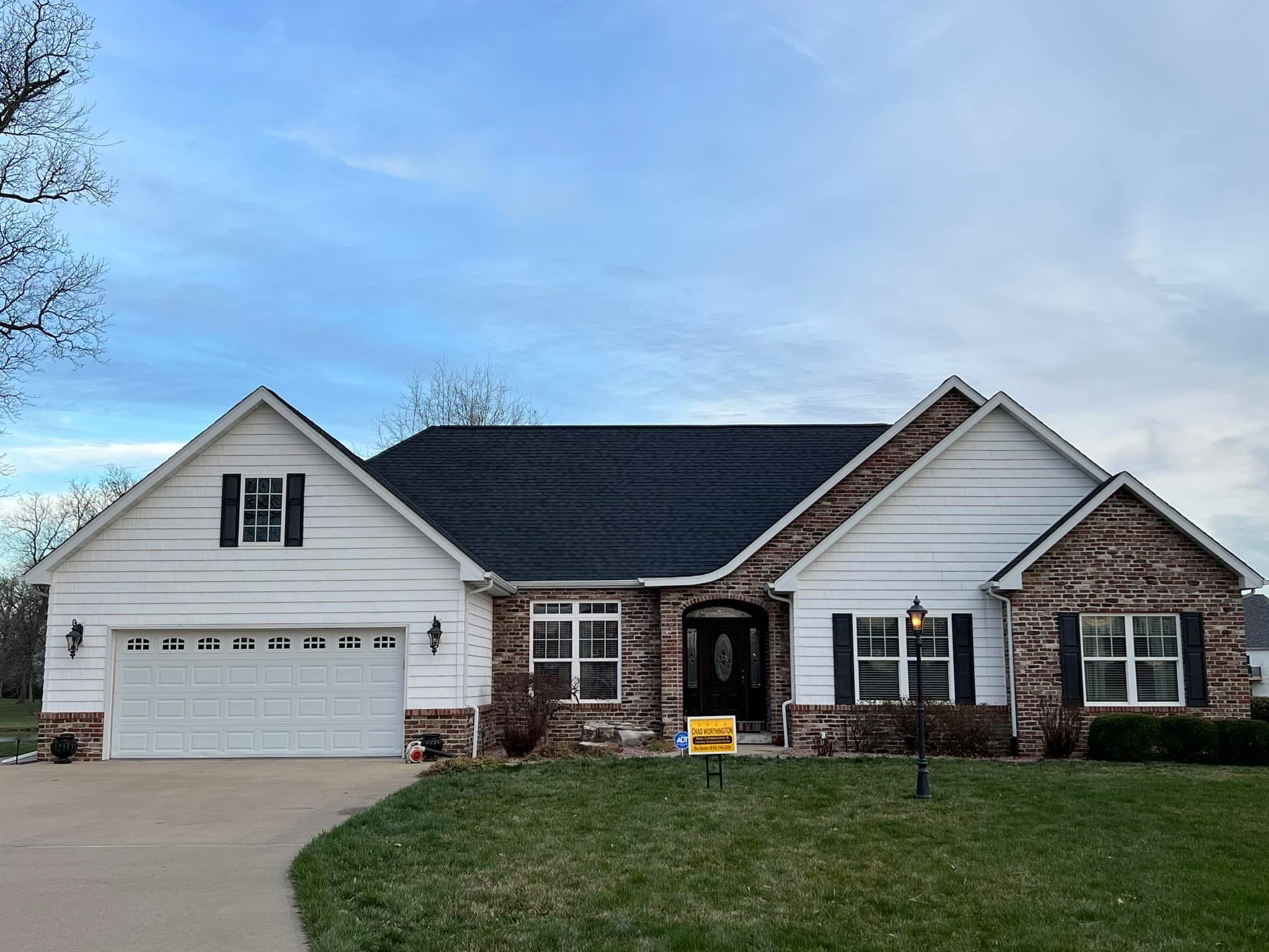 Front view of a two-story house with white siding and a brick facade, black shutters, a garage door, and a front door, with a lawn, sidewalk, and streetlamp in front, and a clear blue sky overhead.