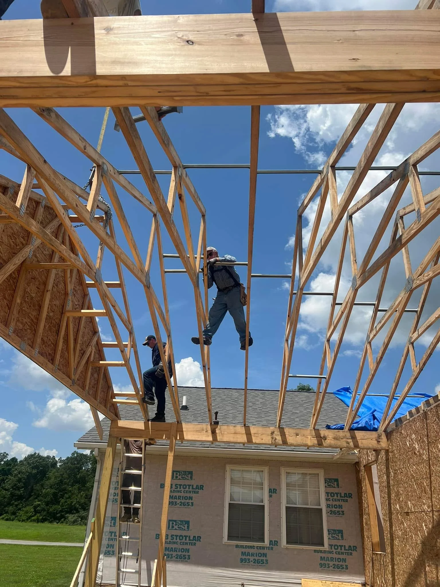 Construction workers building a wooden frame for a house extension beneath a blue sky with clouds, with a residential house in the background.