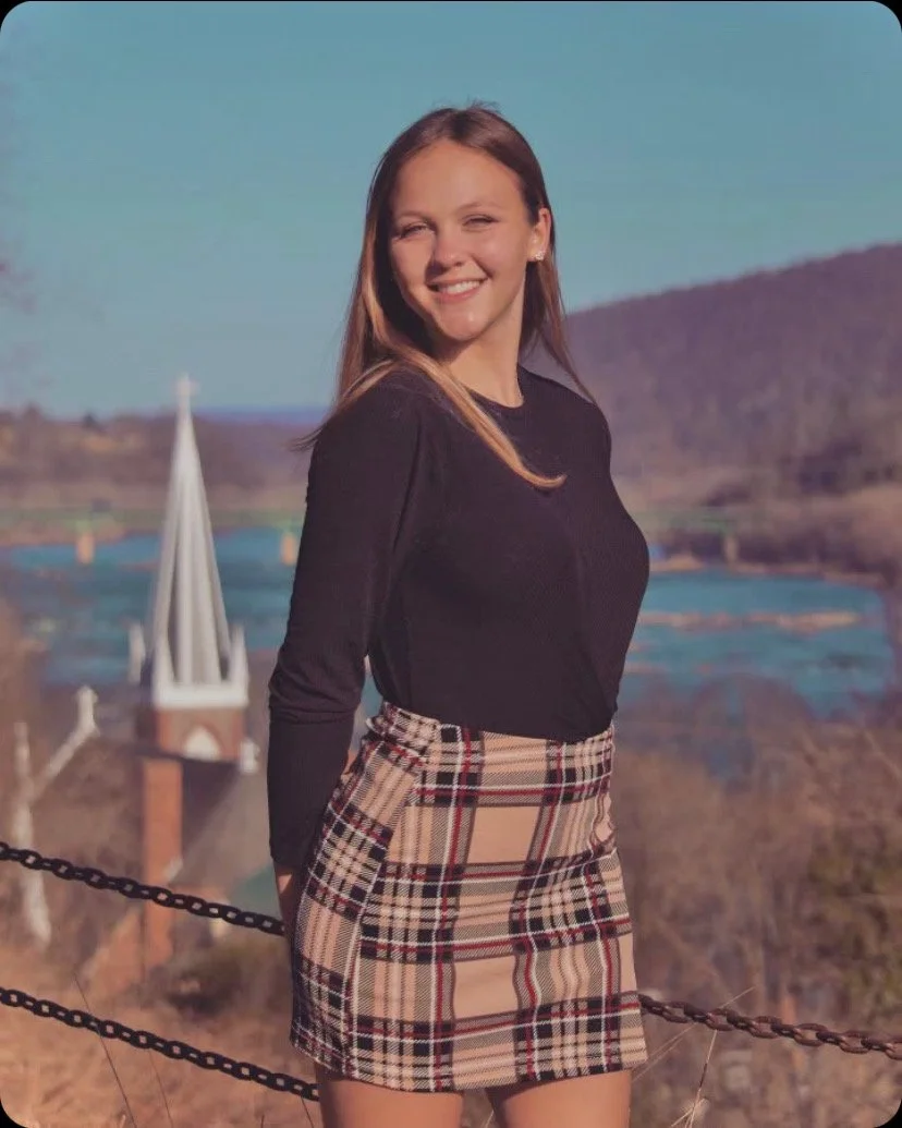 A young woman with long hair smiling outdoors with a river and mountains in the background, wearing a black top and a plaid skirt.