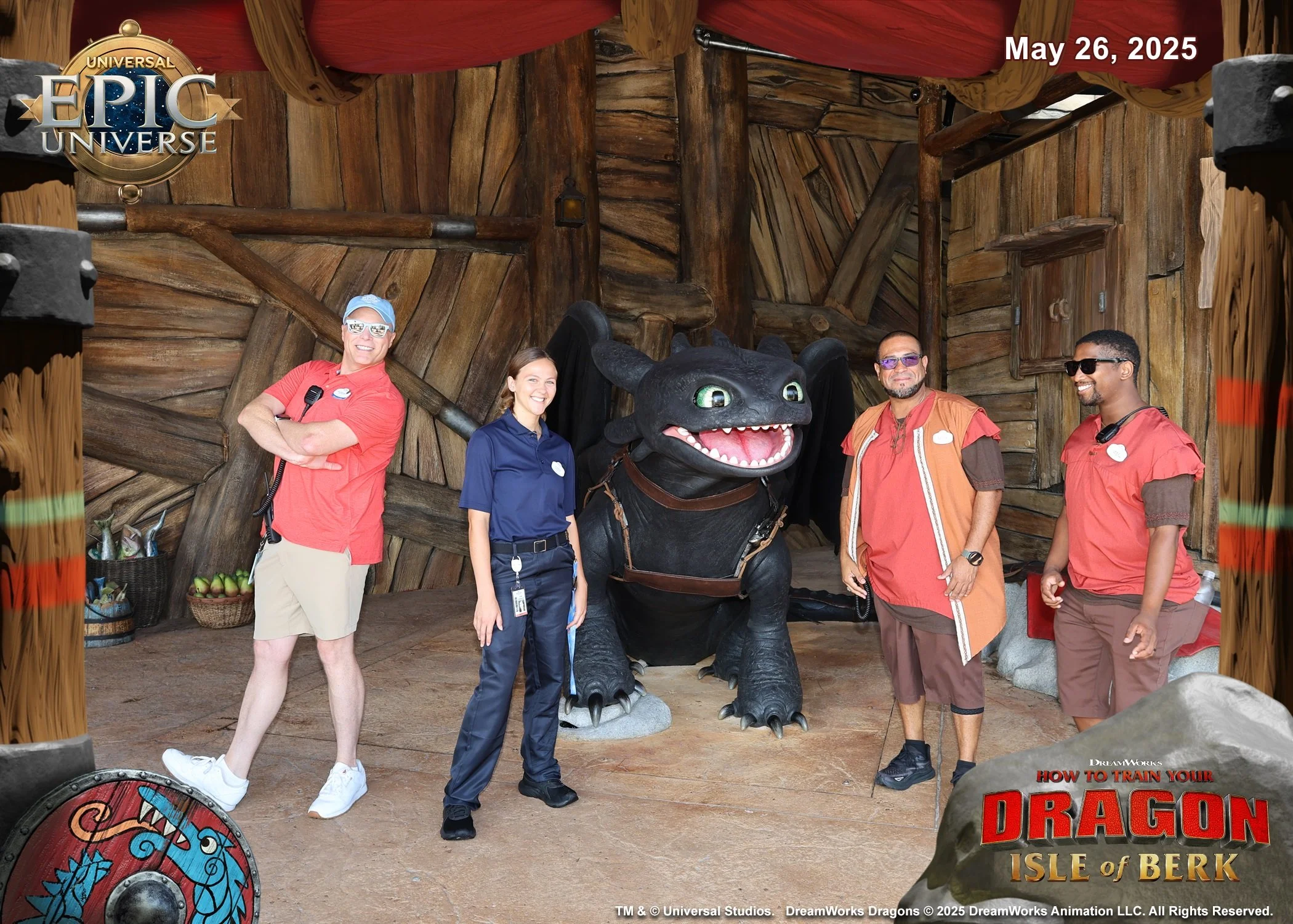 Four people standing in front of a large dragon sculpture inside a themed amusement park attraction with wooden and medieval decor. The photo includes two women and two men, all smiling, with one woman wearing a blue uniform and the others in casual clothing. A dragon head with green eyes and sharp teeth is between them. The background features a wooden wall, lantern, and red canopy. The image has text indicating the date May 26, 2025, and promotional content for a Disney movie, 'How to Train Your Dragon: The Isle of Berk'.