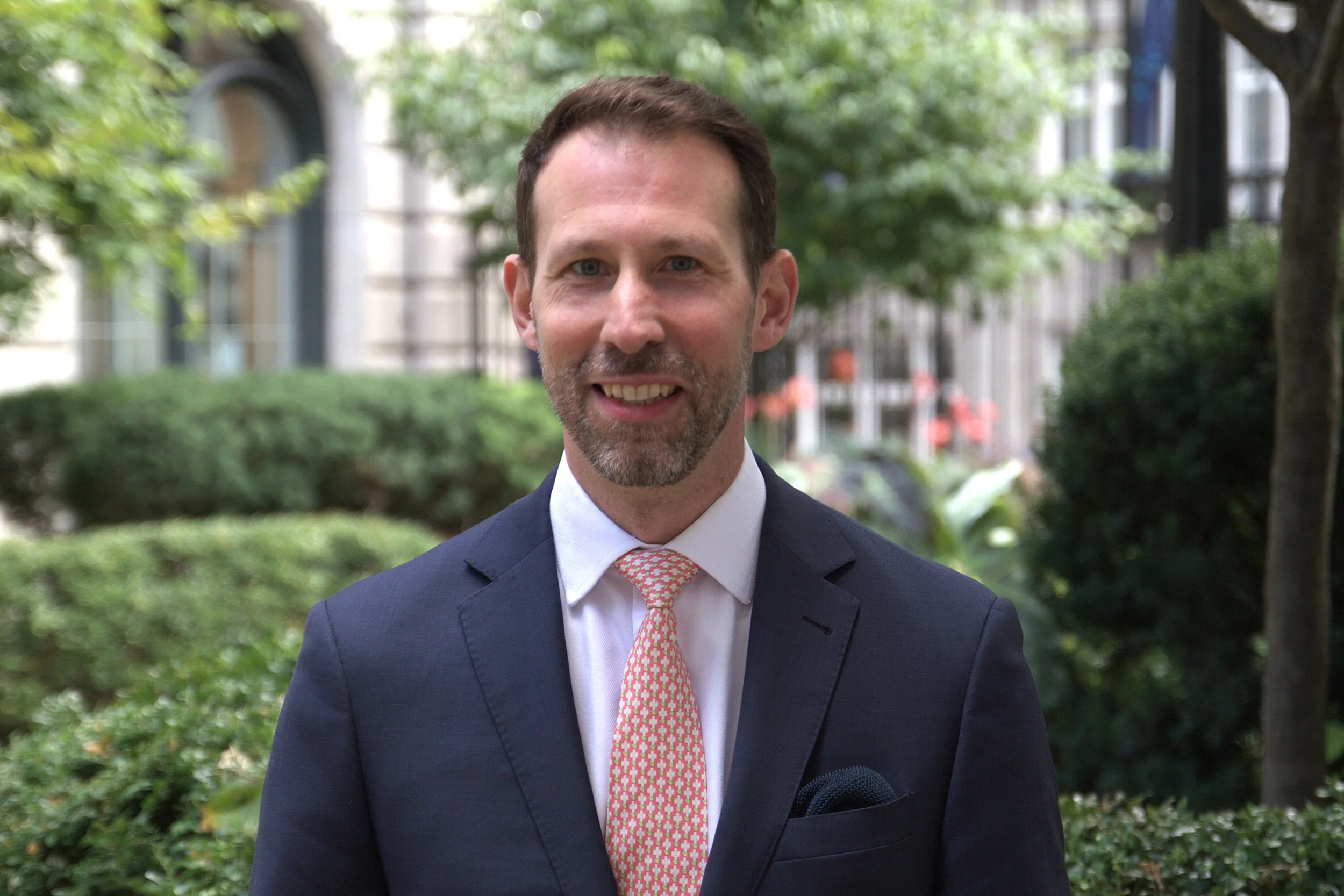 Close-up of a man smiling outdoors, wearing a checkered dress shirt, red tie, and wristwatch, with arms crossed and background of green leaves and buildings.
