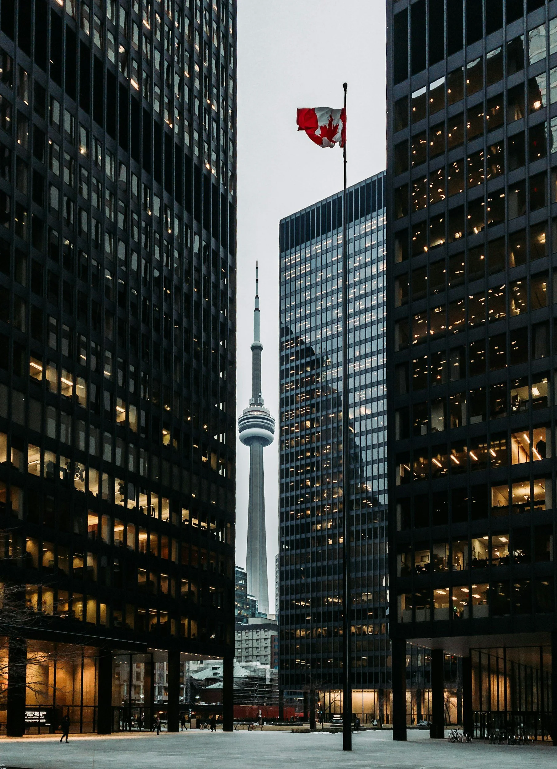 A city scene featuring the Toronto CN Tower visible between two tall office buildings with glass facades at dusk or evening. The buildings frame the tower in the middle, and there are a few pedestrians visible on the sidewalk, along with some bicycles.