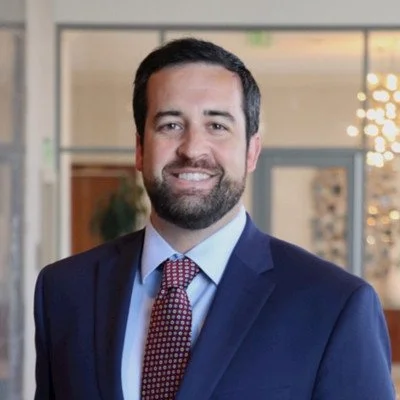 A man in a navy suit, light blue shirt, and patterned tie smiling, standing in a modern office environment.